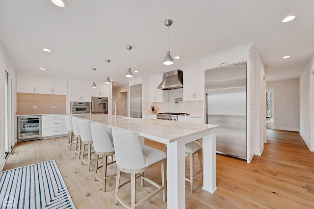 kitchen with light wood-style flooring, stainless steel appliances, white cabinets, wine cooler, and wall chimney range hood