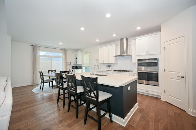 kitchen featuring stainless steel double oven, a sink, gas stovetop, wall chimney exhaust hood, and a center island with sink