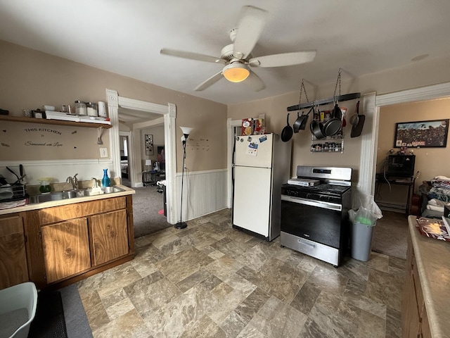 kitchen with wainscoting, brown cabinets, freestanding refrigerator, stainless steel gas range, and a sink