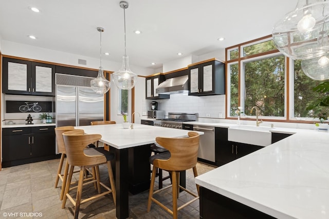 kitchen with dark cabinets, a sink, appliances with stainless steel finishes, wall chimney exhaust hood, and tasteful backsplash