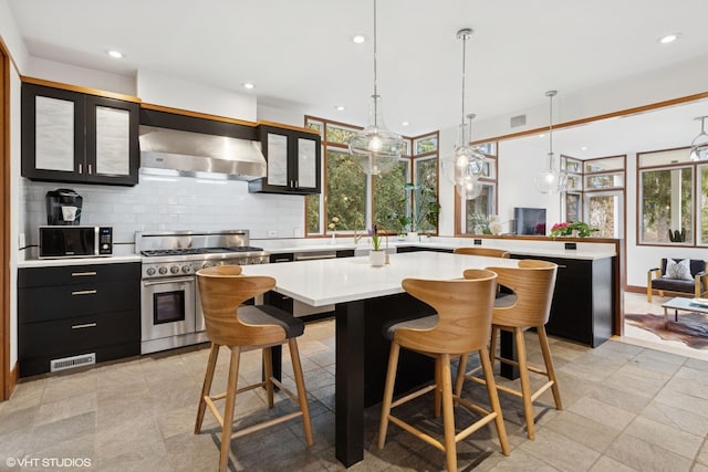 kitchen featuring range with two ovens, light countertops, wall chimney range hood, dark cabinetry, and tasteful backsplash