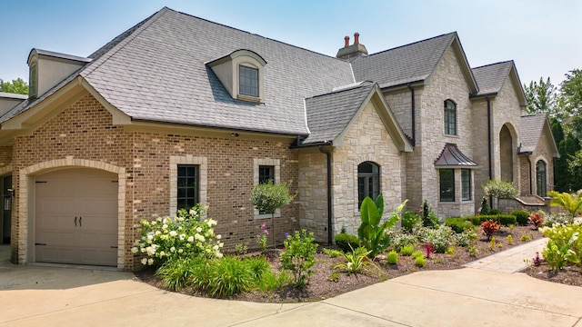 french provincial home featuring driveway, brick siding, an attached garage, and a high end roof