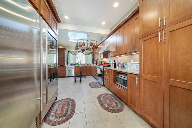 kitchen featuring light tile patterned flooring, under cabinet range hood, a skylight, appliances with stainless steel finishes, and brown cabinetry