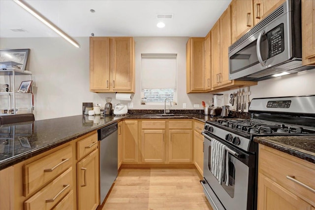 kitchen featuring light brown cabinets, a sink, appliances with stainless steel finishes, light wood finished floors, and dark stone countertops