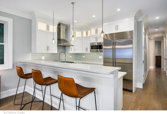 kitchen featuring built in appliances, a peninsula, wall chimney range hood, and white cabinetry