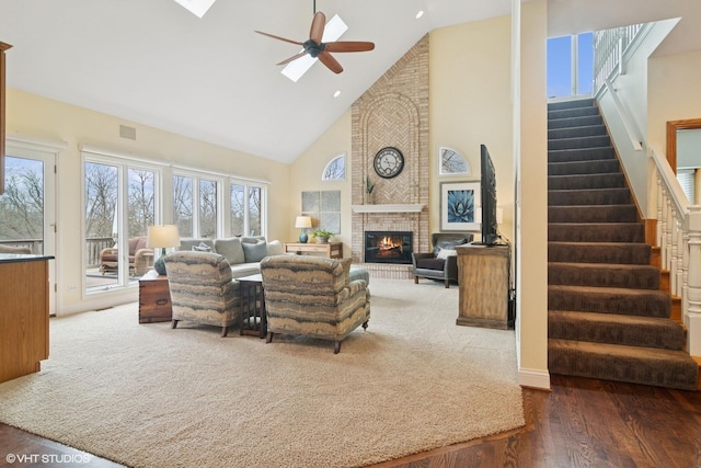 carpeted living room with high vaulted ceiling, stairway, a ceiling fan, a brick fireplace, and visible vents