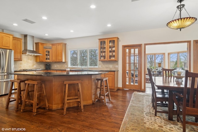 kitchen with freestanding refrigerator, a kitchen island, dark wood-style floors, wall chimney range hood, and glass insert cabinets