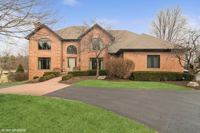 french country home with a chimney, brick siding, aphalt driveway, a front lawn, and a shingled roof
