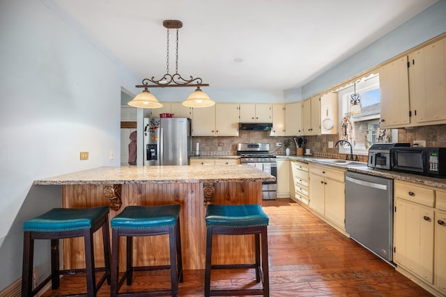 kitchen featuring a sink, appliances with stainless steel finishes, cream cabinetry, a peninsula, and under cabinet range hood