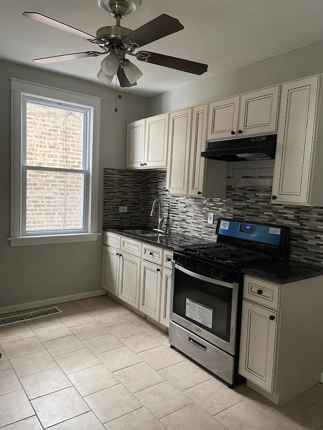 kitchen with dark countertops, under cabinet range hood, cream cabinets, stainless steel gas stove, and a sink