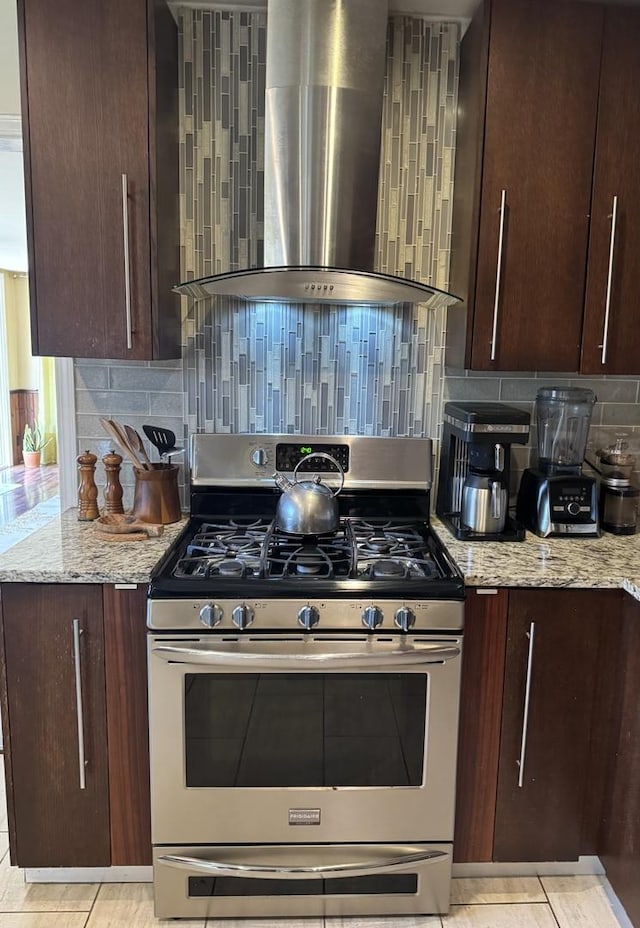 kitchen with stainless steel gas stove, dark brown cabinetry, wall chimney exhaust hood, and light stone countertops