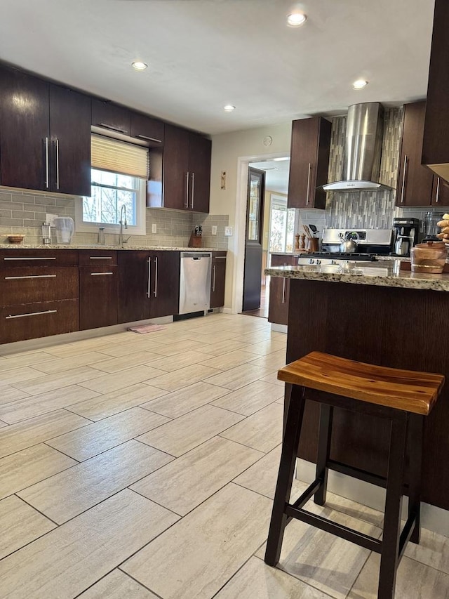 kitchen featuring dark brown cabinetry, light stone counters, wall chimney exhaust hood, and appliances with stainless steel finishes