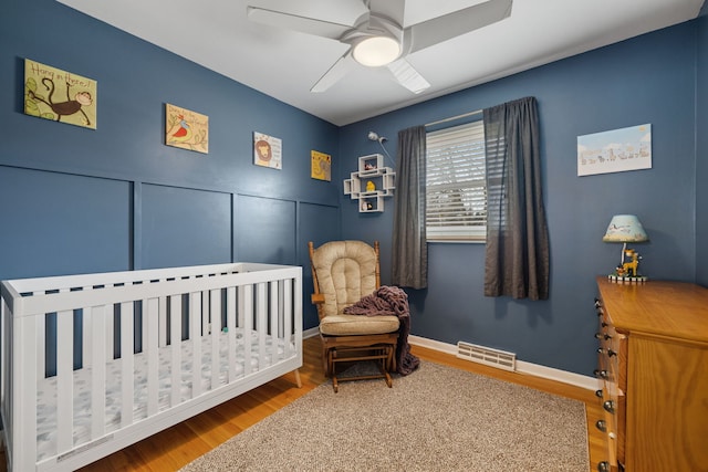 bedroom with wood finished floors, visible vents, baseboards, a ceiling fan, and a nursery area