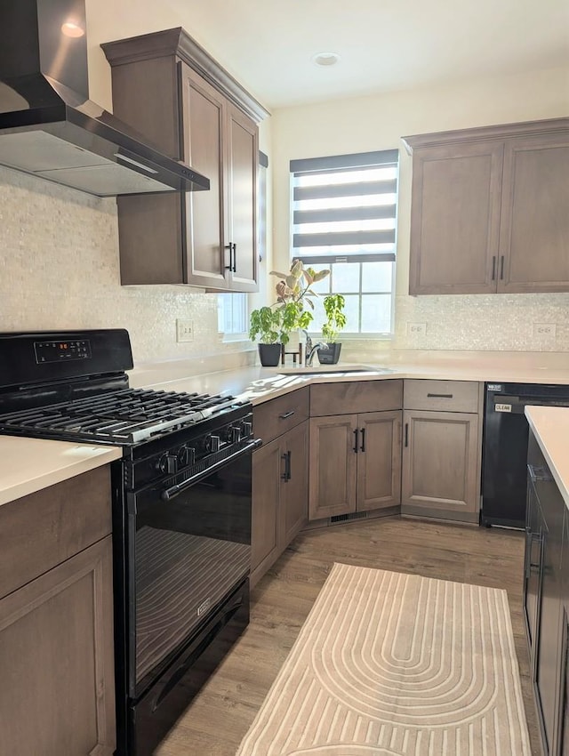 kitchen featuring black appliances, light wood-style flooring, wall chimney range hood, backsplash, and light countertops