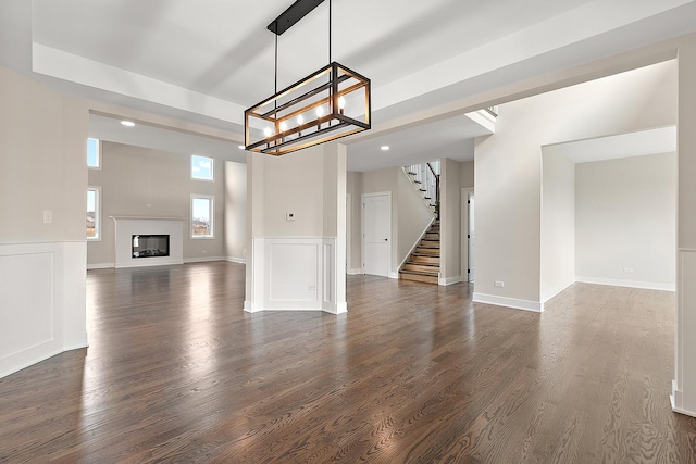 unfurnished living room with dark wood finished floors, a decorative wall, stairway, a glass covered fireplace, and wainscoting