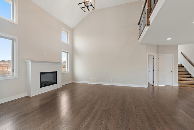 unfurnished living room with dark wood-type flooring, a glass covered fireplace, high vaulted ceiling, baseboards, and stairs