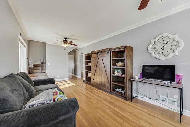 living room featuring light wood finished floors, a barn door, baseboards, ceiling fan, and crown molding