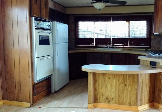 kitchen featuring light wood-style flooring, a sink, wooden walls, ceiling fan, and white appliances