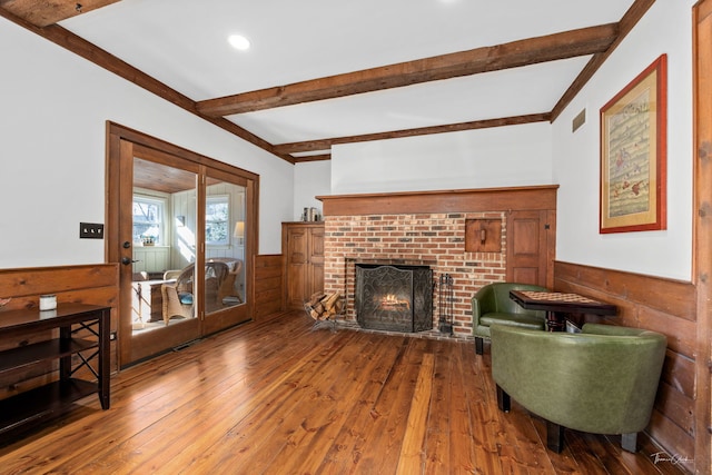 living room featuring a wainscoted wall, wood-type flooring, visible vents, a brick fireplace, and beamed ceiling