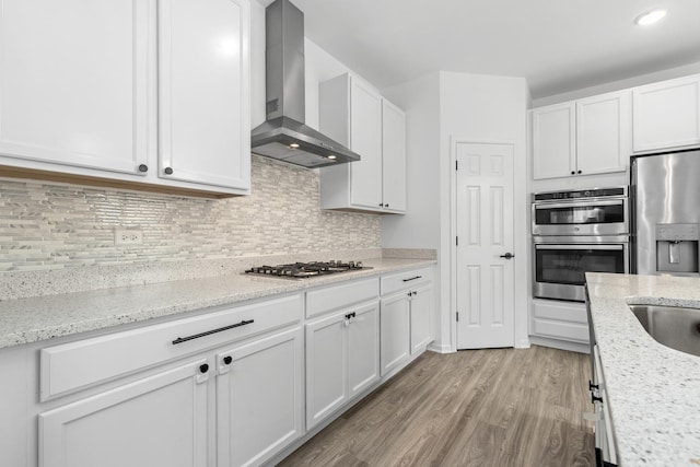 kitchen featuring light wood finished floors, tasteful backsplash, wall chimney exhaust hood, appliances with stainless steel finishes, and white cabinetry