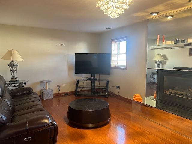 living area with visible vents, baseboards, a glass covered fireplace, wood finished floors, and a notable chandelier