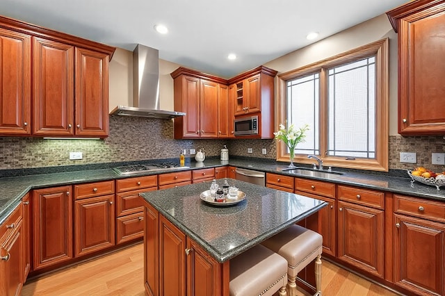 kitchen with a breakfast bar area, a sink, light wood-style floors, appliances with stainless steel finishes, and wall chimney exhaust hood