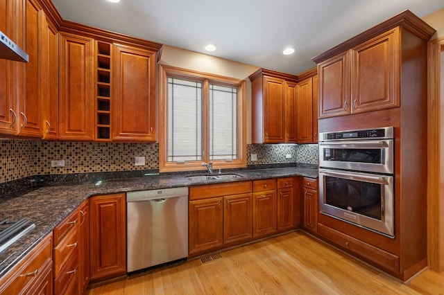 kitchen featuring light wood finished floors, tasteful backsplash, appliances with stainless steel finishes, a sink, and dark stone counters