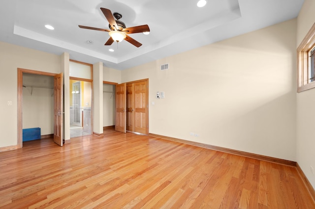 unfurnished bedroom featuring multiple closets, a tray ceiling, light wood-type flooring, and baseboards