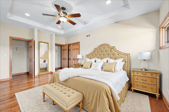 bedroom with baseboards, visible vents, wood finished floors, a tray ceiling, and multiple closets