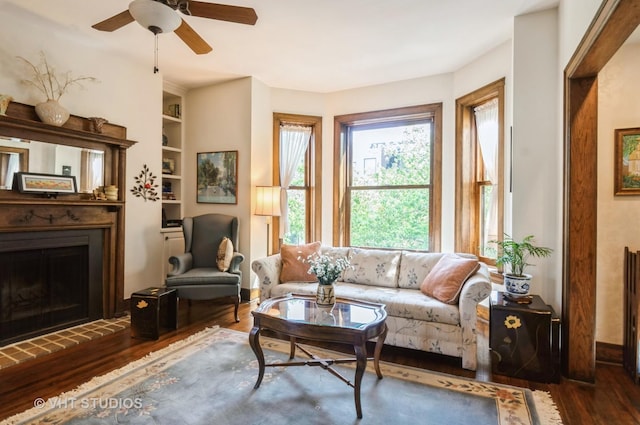 living room with built in shelves, a ceiling fan, a fireplace with raised hearth, and wood finished floors