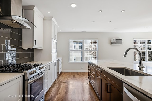 kitchen with light stone countertops, a sink, stainless steel appliances, wall chimney range hood, and tasteful backsplash