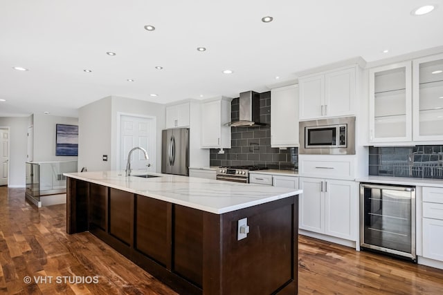 kitchen featuring an island with sink, beverage cooler, a sink, stainless steel appliances, and wall chimney range hood