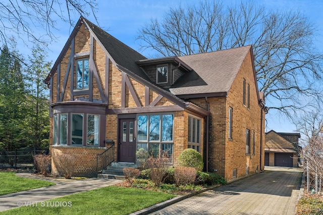 english style home featuring an outbuilding, fence, a shingled roof, a garage, and brick siding