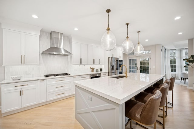 kitchen with an island with sink, a sink, appliances with stainless steel finishes, wall chimney range hood, and decorative backsplash