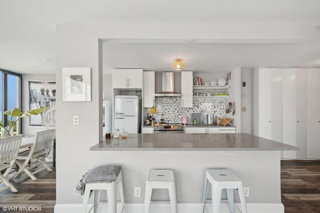 kitchen with stainless steel electric stove, freestanding refrigerator, white cabinetry, a peninsula, and wall chimney exhaust hood