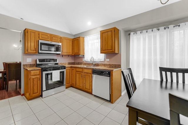 kitchen featuring stainless steel appliances, brown cabinetry, light tile patterned flooring, a sink, and light stone countertops