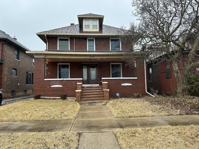 american foursquare style home with covered porch, a shingled roof, and brick siding