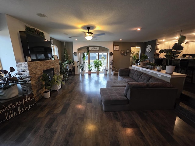 living area featuring ceiling fan, a textured ceiling, a stone fireplace, arched walkways, and dark wood-style floors