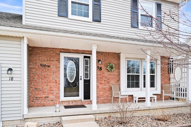 doorway to property with covered porch, a shingled roof, and brick siding