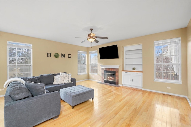 living room featuring a ceiling fan, baseboards, built in features, light wood-style floors, and a brick fireplace