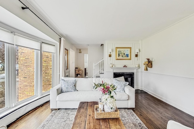 living room featuring a glass covered fireplace, wood finished floors, a baseboard radiator, baseboards, and stairs