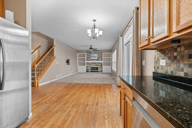 living room with stairs, visible vents, light wood-style flooring, a stone fireplace, and ceiling fan with notable chandelier