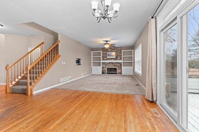 unfurnished living room featuring stairs, visible vents, a stone fireplace, and ceiling fan with notable chandelier