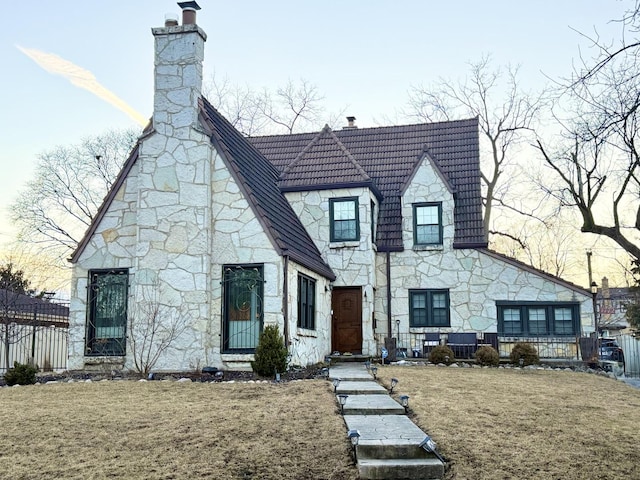 view of front of property featuring stone siding, a chimney, a tiled roof, fence, and a front lawn