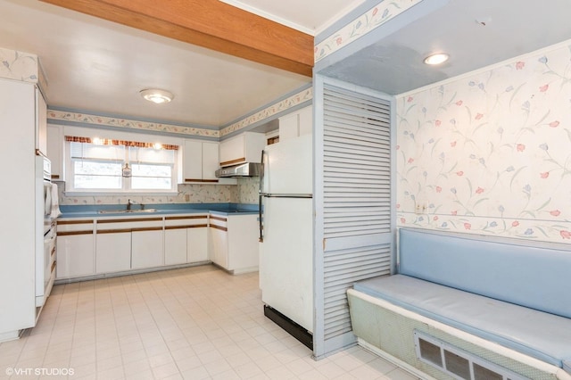 kitchen with a sink, under cabinet range hood, white appliances, white cabinets, and wallpapered walls