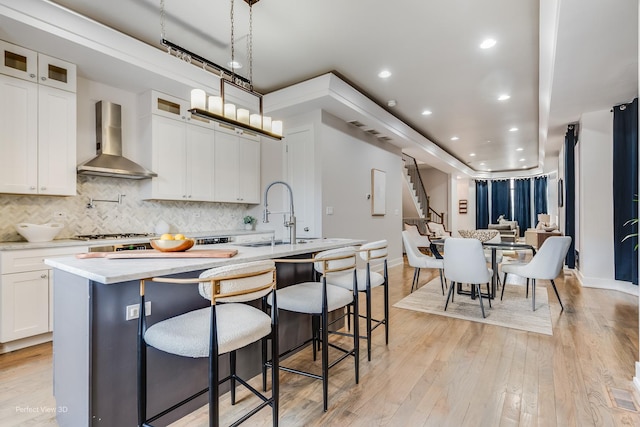 kitchen featuring a center island with sink, light wood-style flooring, backsplash, a sink, and wall chimney exhaust hood