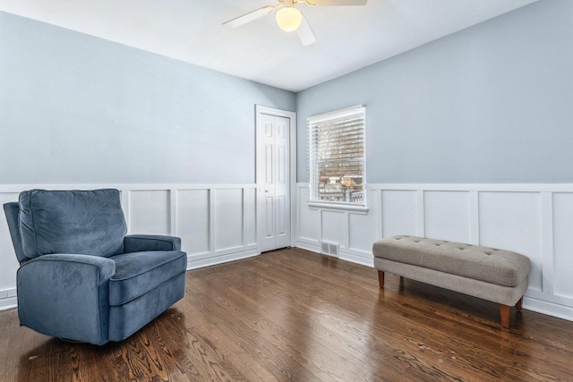 sitting room with ceiling fan, visible vents, dark wood-style floors, and a decorative wall