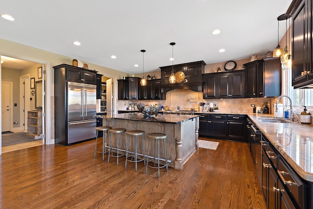 kitchen with dark stone countertops, dark wood-type flooring, a center island, stainless steel built in fridge, and a sink