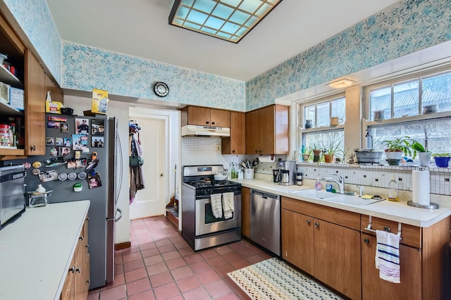 kitchen with wallpapered walls, brown cabinetry, stainless steel appliances, under cabinet range hood, and a sink