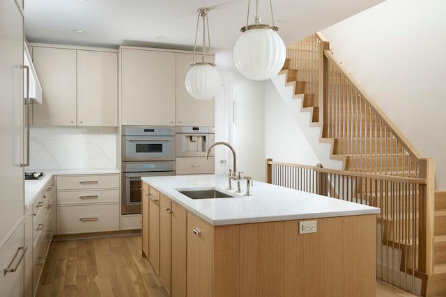 kitchen with a kitchen island with sink, decorative light fixtures, a sink, double oven, and light wood finished floors
